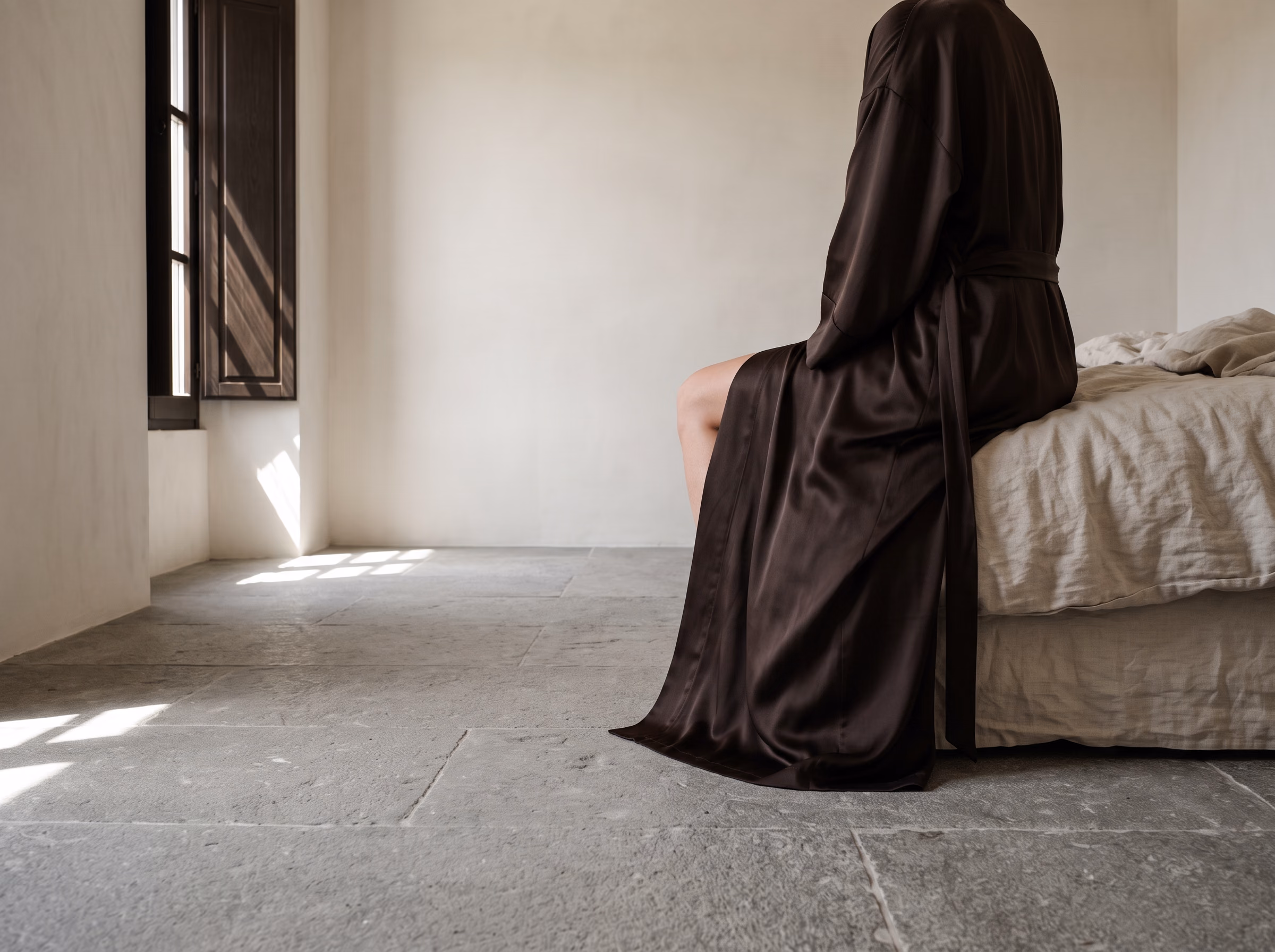 The hem of a Noyau robe, seated at the edge of the bed. Shutters cut the light across the floor.